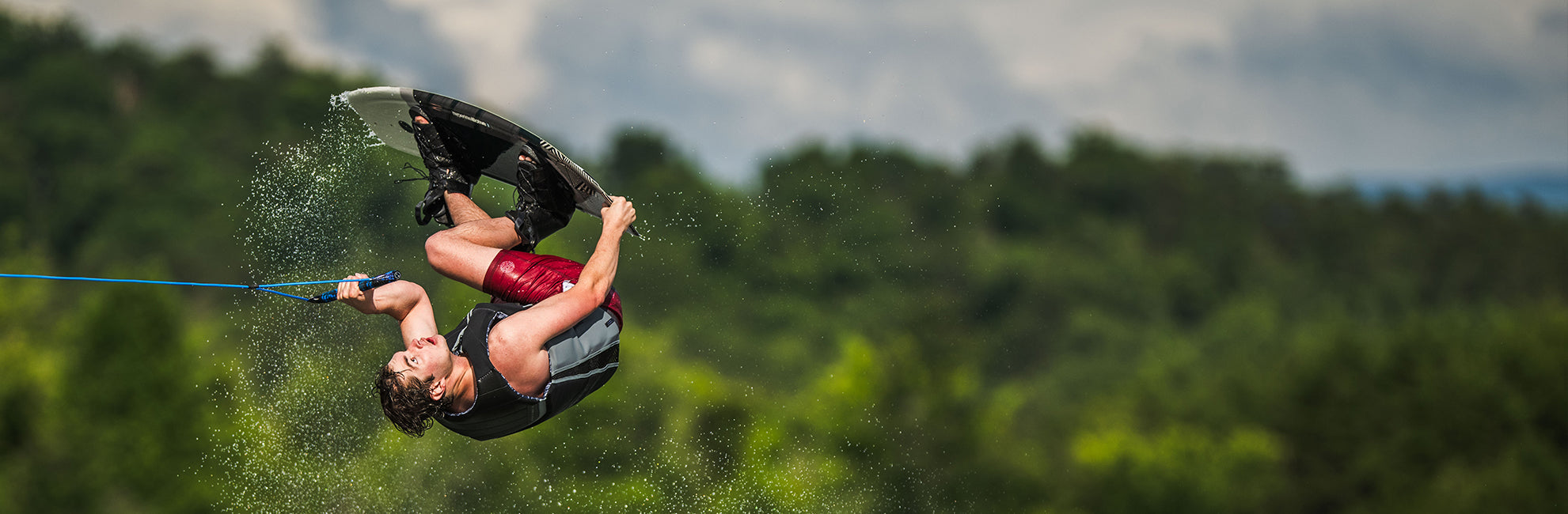 Person wakeboarding in mid-air, performing a flip against a backdrop of trees and a cloudy sky.