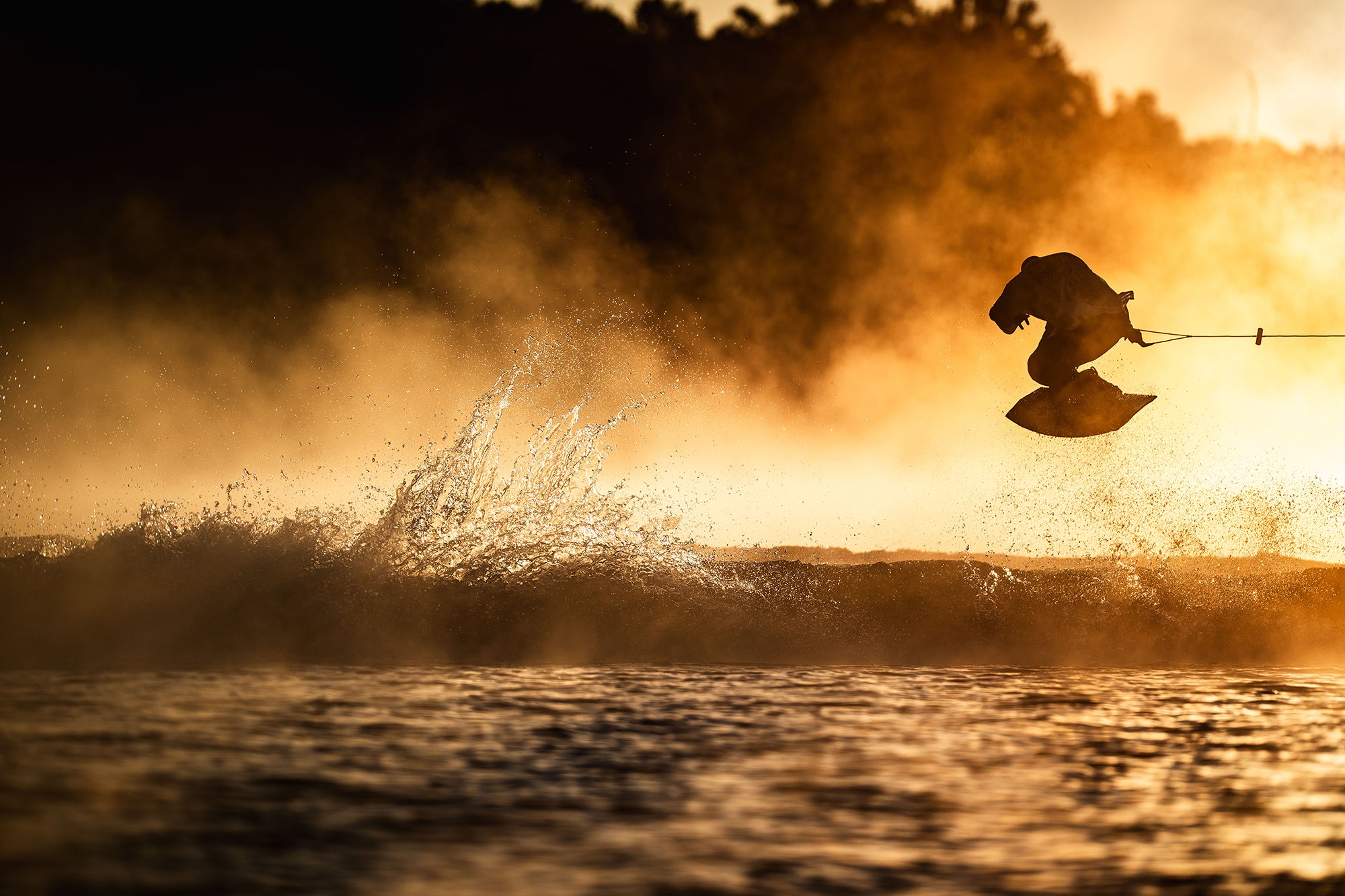 Silhouetted person wakeboarding, catching air at sunset, with water spray and a golden sky in the background.
