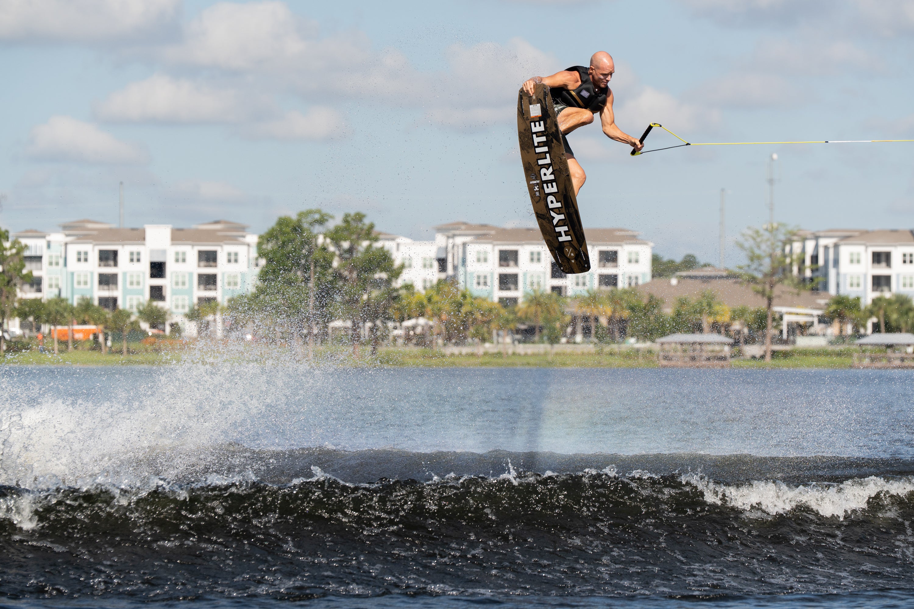 Wakeboarder catches air over the wake in front of lakeside apartments and cloudy sky.