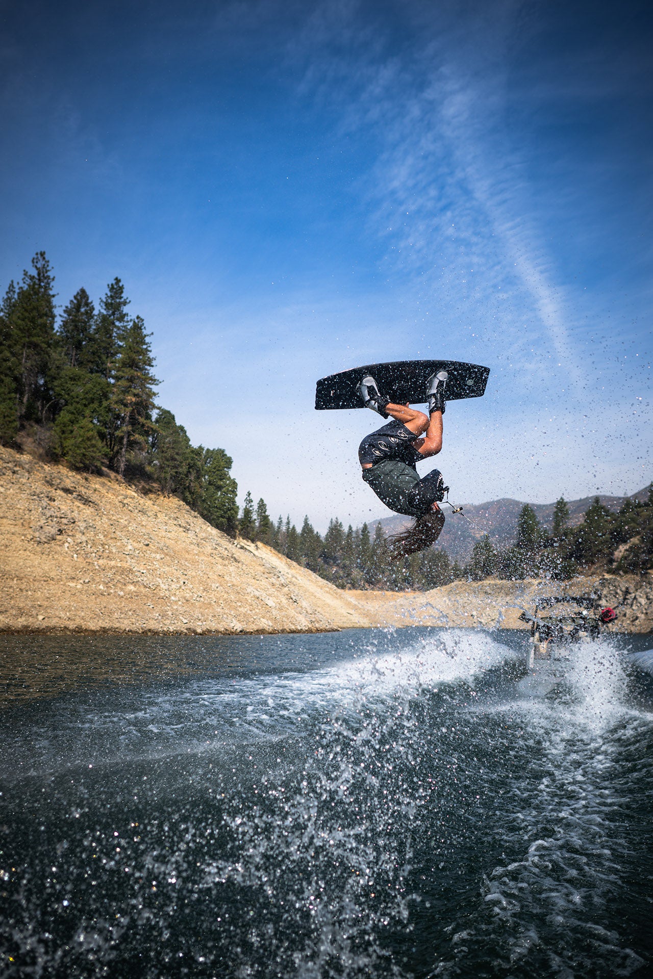 Person wakeboarding, performing a backflip over a lake with trees in the background.