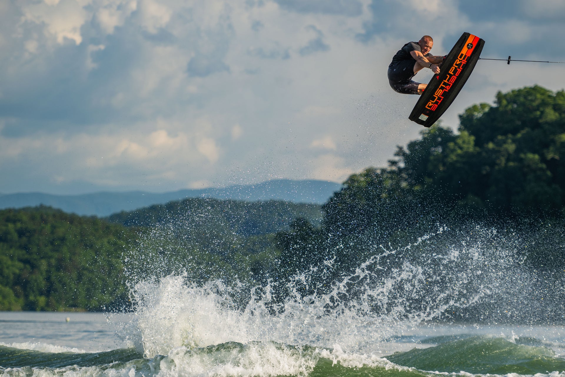 Person wakeboarding in the air over a lake with trees and clouds in the background.