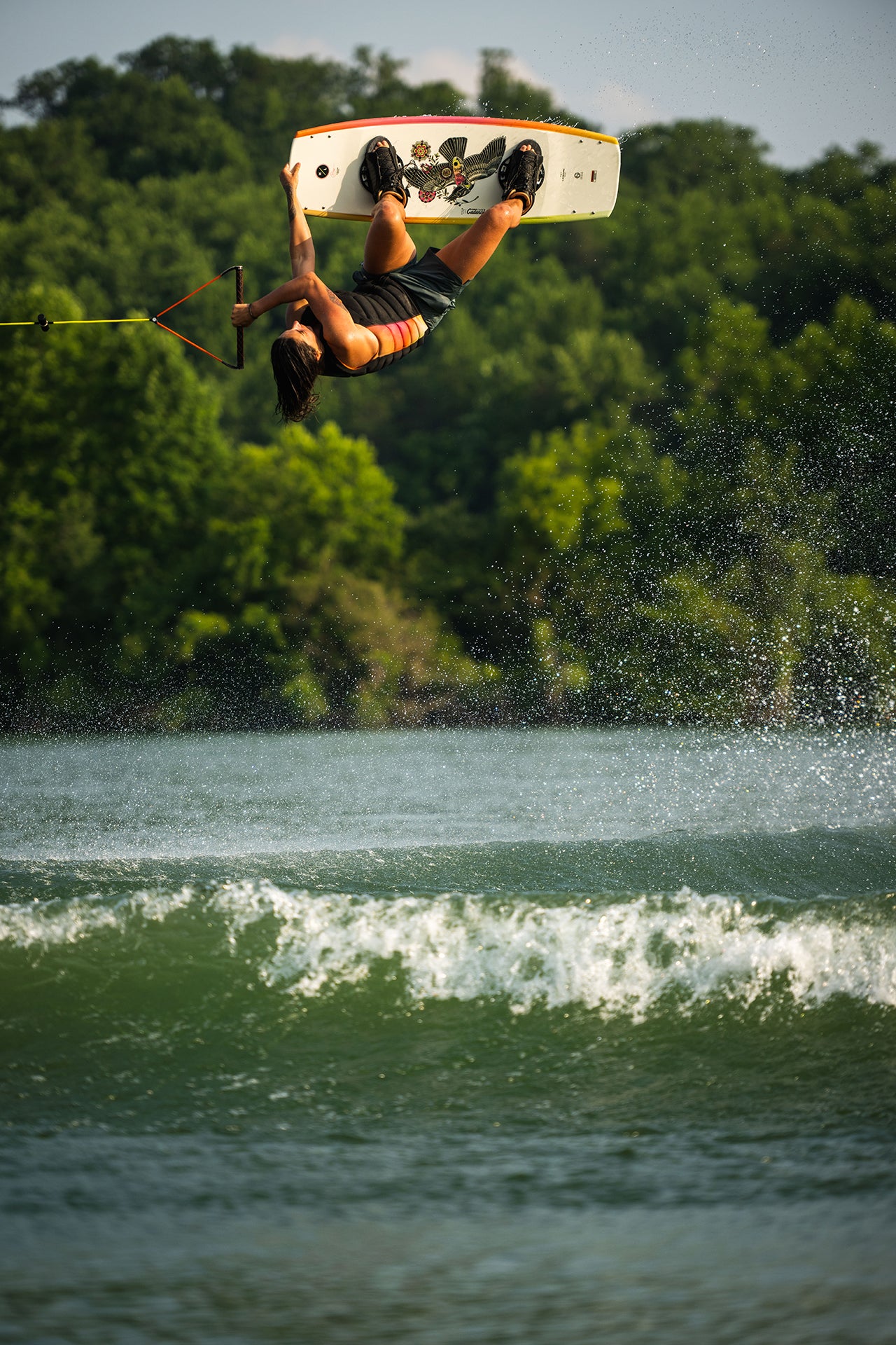 Person wakeboarding and performing a flip over water with trees in the background.
