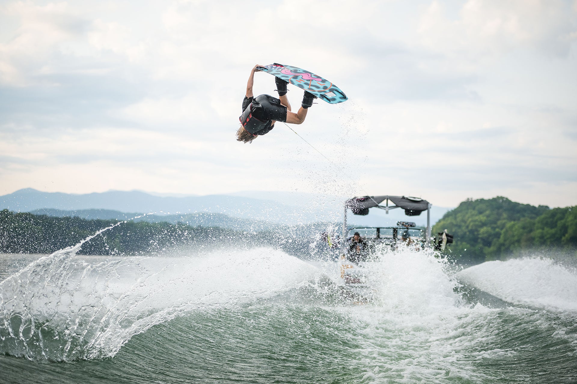 Person wakeboarding, performing a flip over waves with a boat in the backdrop.