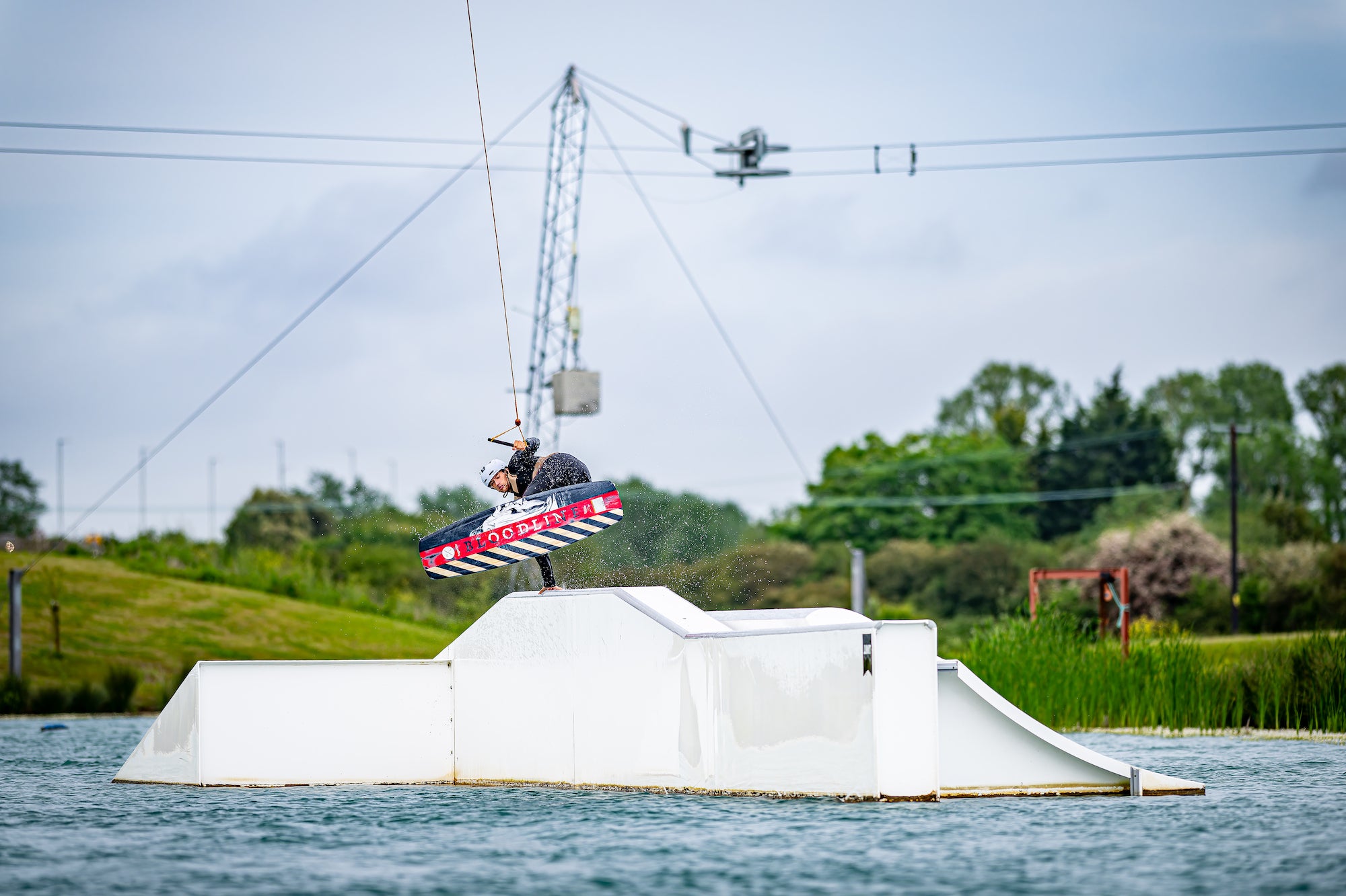 Person wakeboarding over a white ramp on a lake, surrounded by greenery and a cable system overhead.