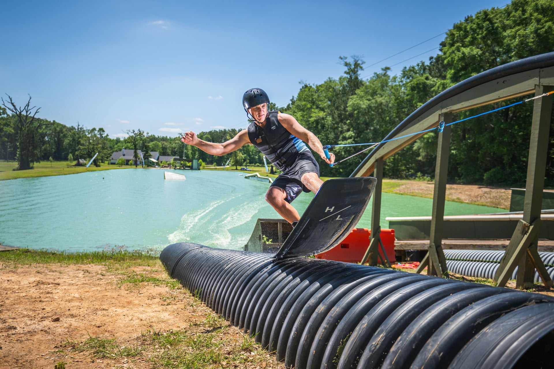 Person wakeboarding on a pipe at a cable park with trees and water in the background.