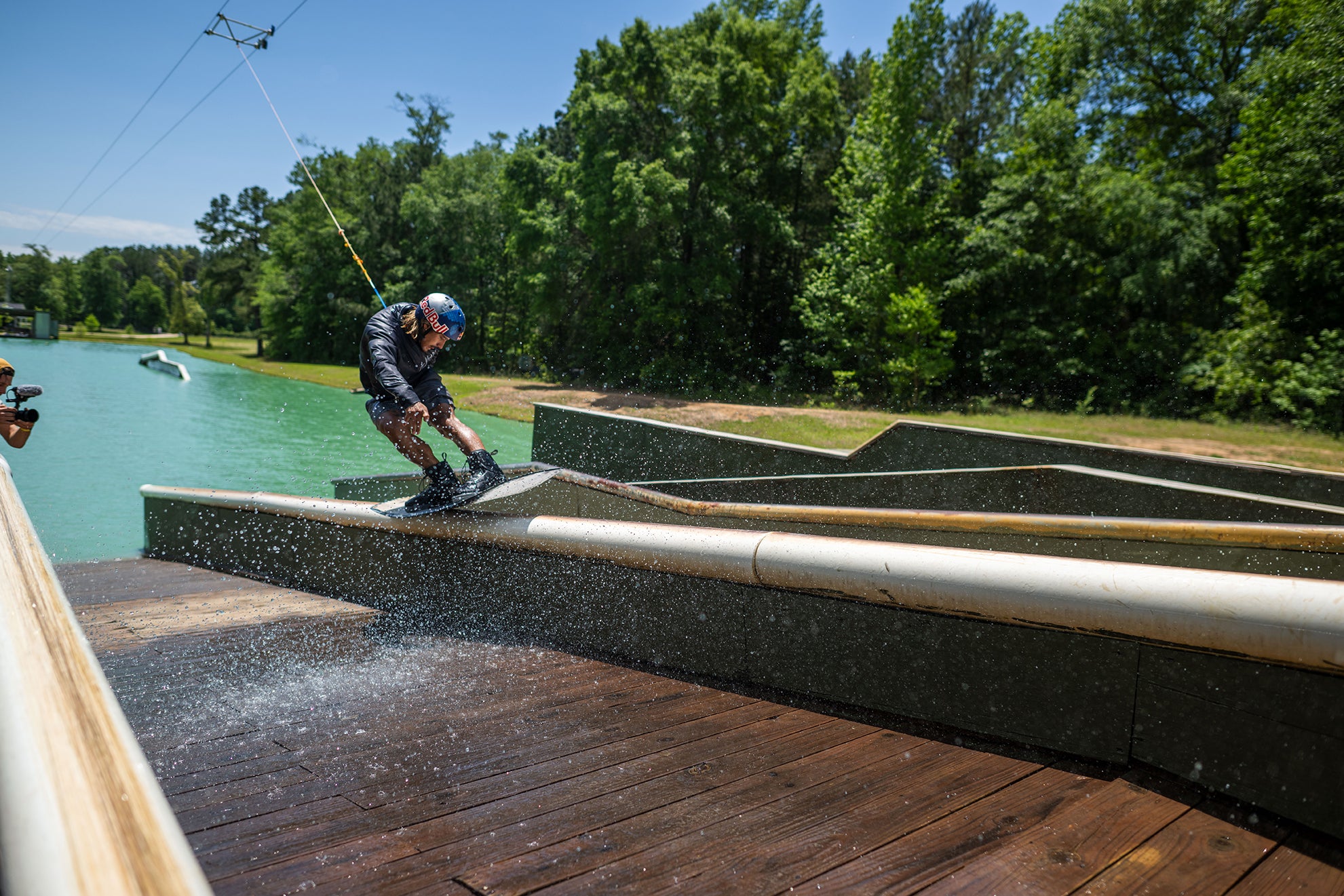 Person wakeboarding on a lake, performing a trick on a wooden ramp with greenery in the background.