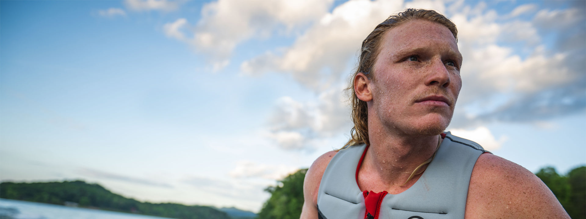 A person with long hair wearing a life vest gazes thoughtfully over a lake, with trees and a cloudy sky in the background.
