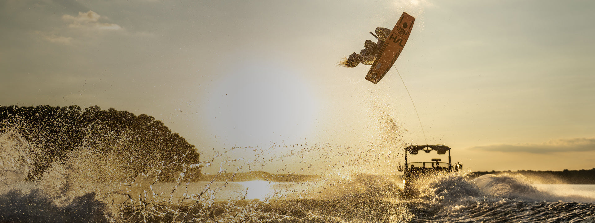 A person wakeboarding and catching air behind a boat on a sunlit lake, with splashing water.