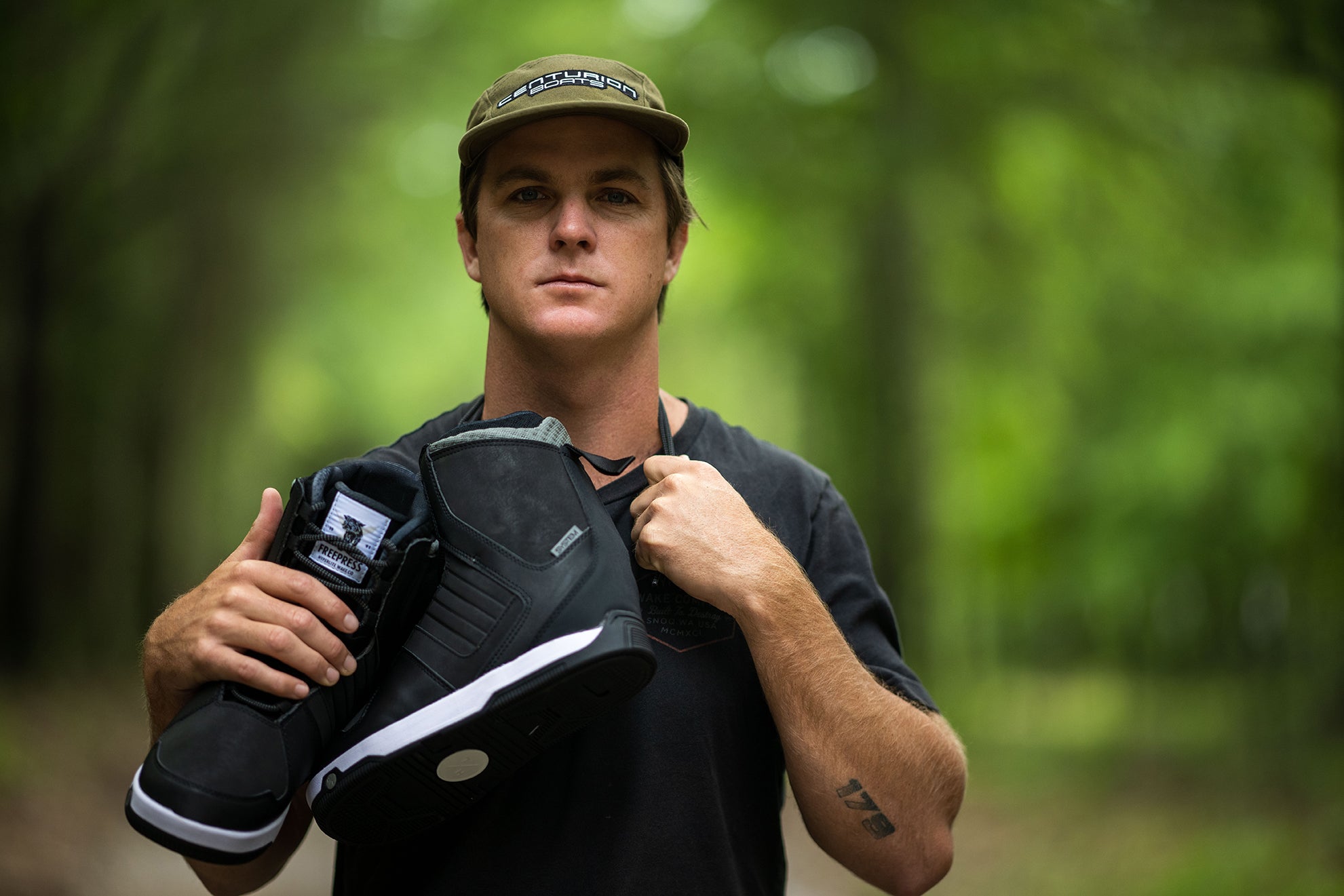 Person wearing a black t-shirt and cap, holding black boots over their shoulder in a forest setting.