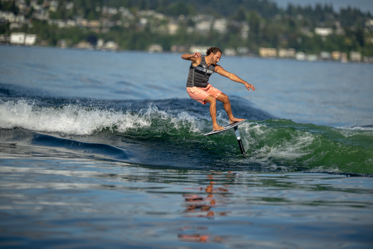 Person riding a hydrofoil surfboard, skimming over water with a forested shoreline in the background.