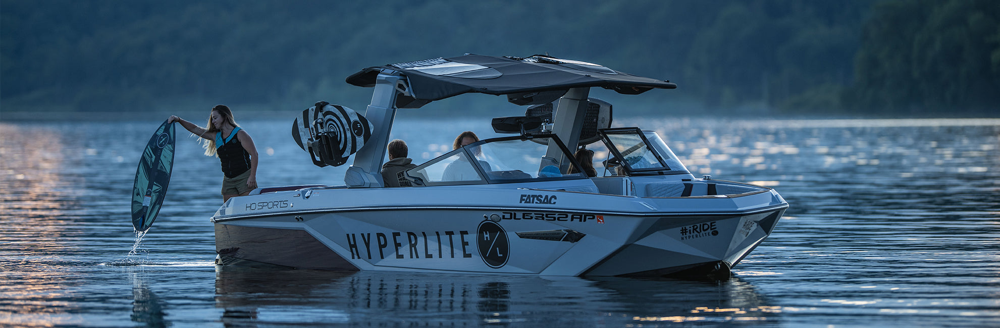 A wakeboarding boat on a lake with people and gear, surrounded by a wooded shoreline at dusk.