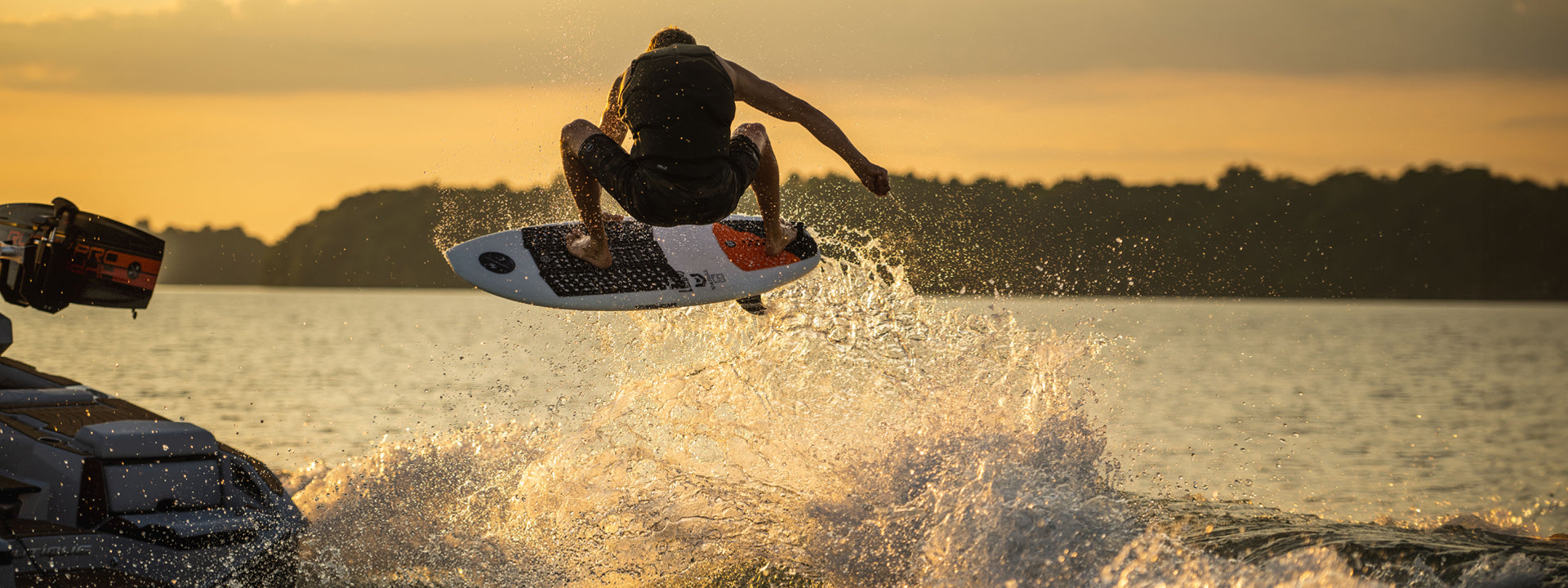 A person wakeboarding on a lake at sunset, performing a jump with water splashing around.