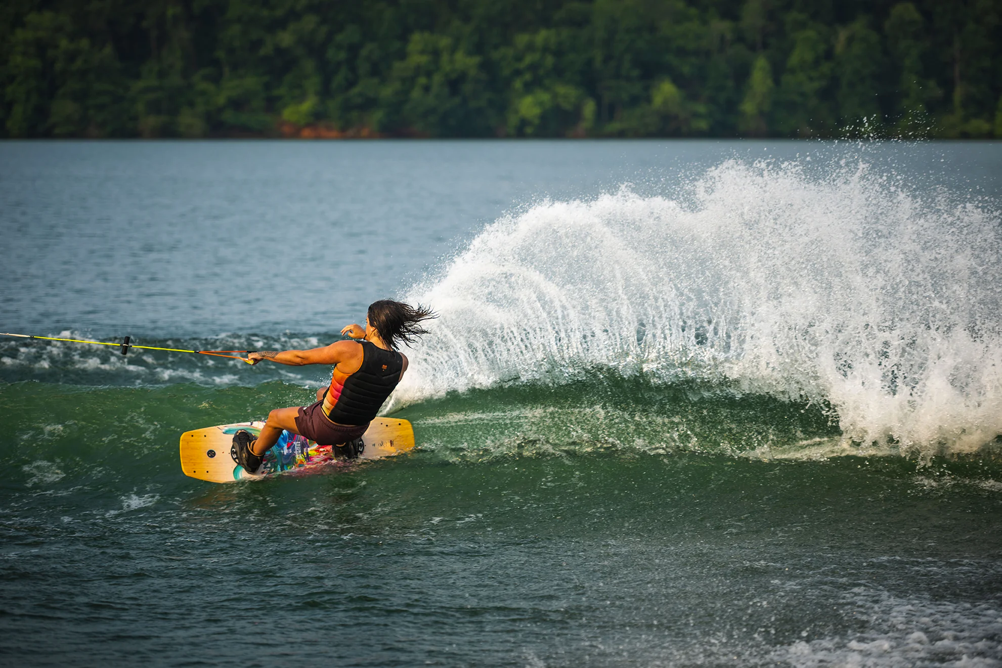 Person wakeboarding on a lake, creating a large spray of water, with a forested backdrop.