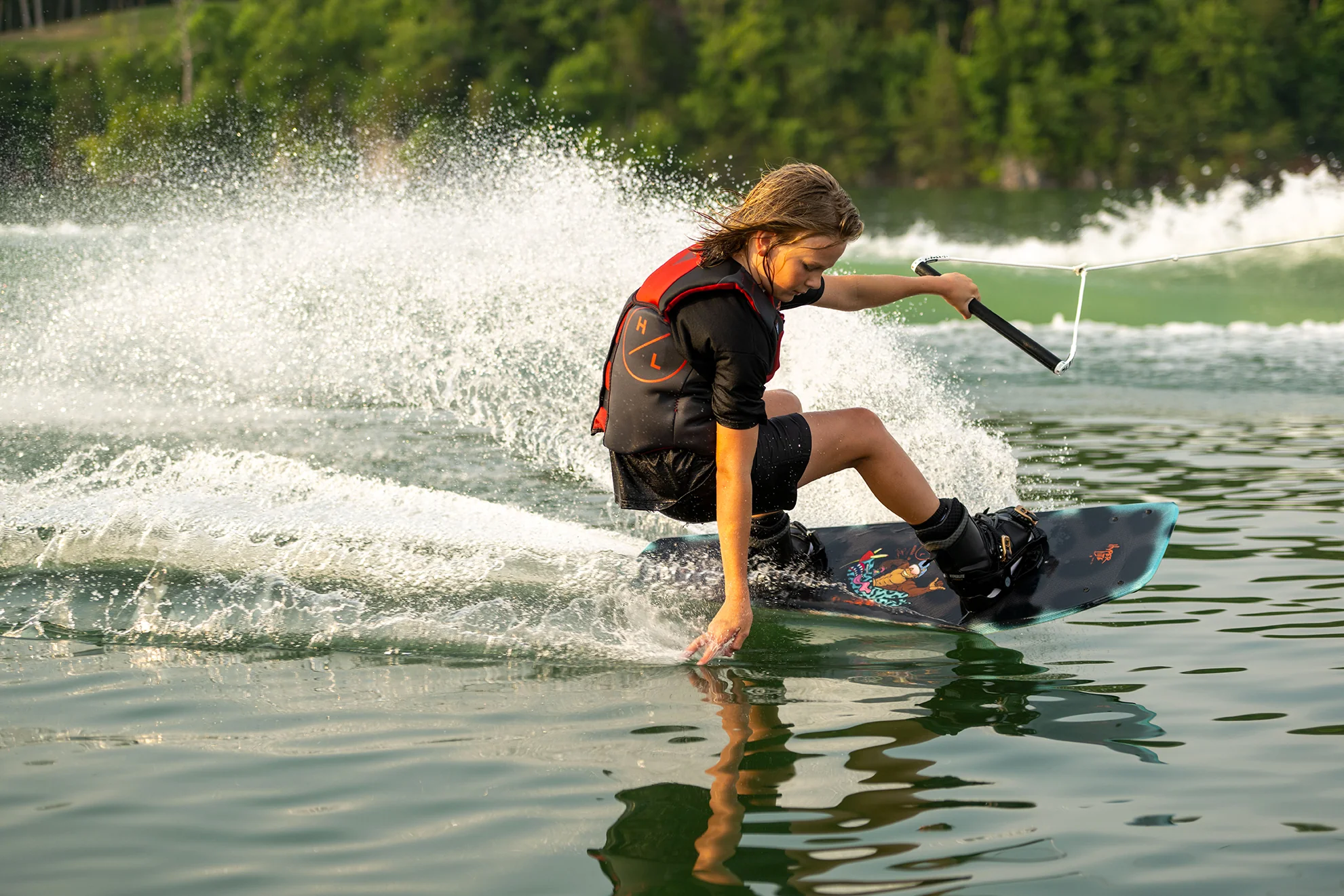 Person wakeboarding on a lake, leaning back and creating a splash, with green trees in the background.