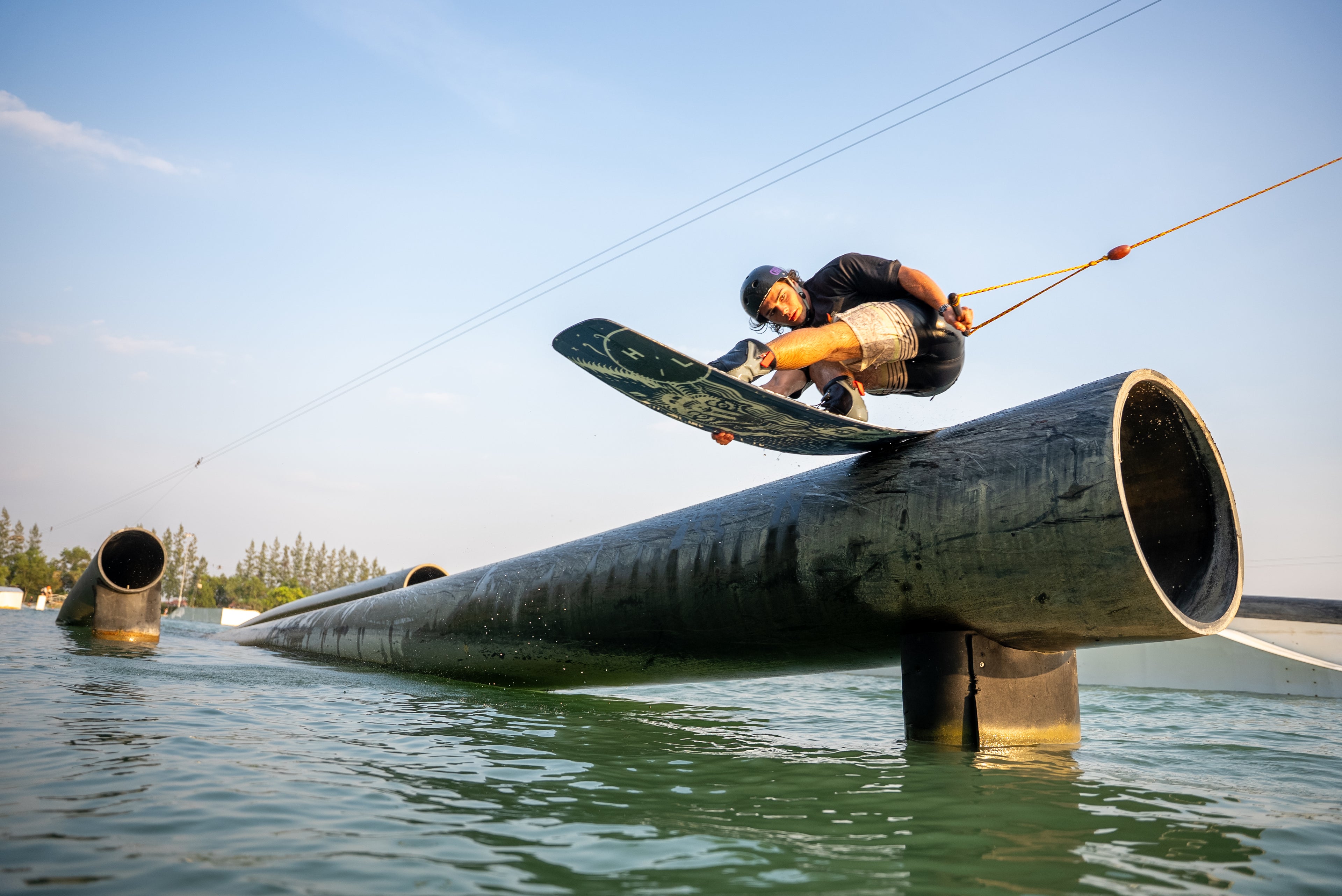 Person wakeboarding over a pipe on water, with a clear sky background.