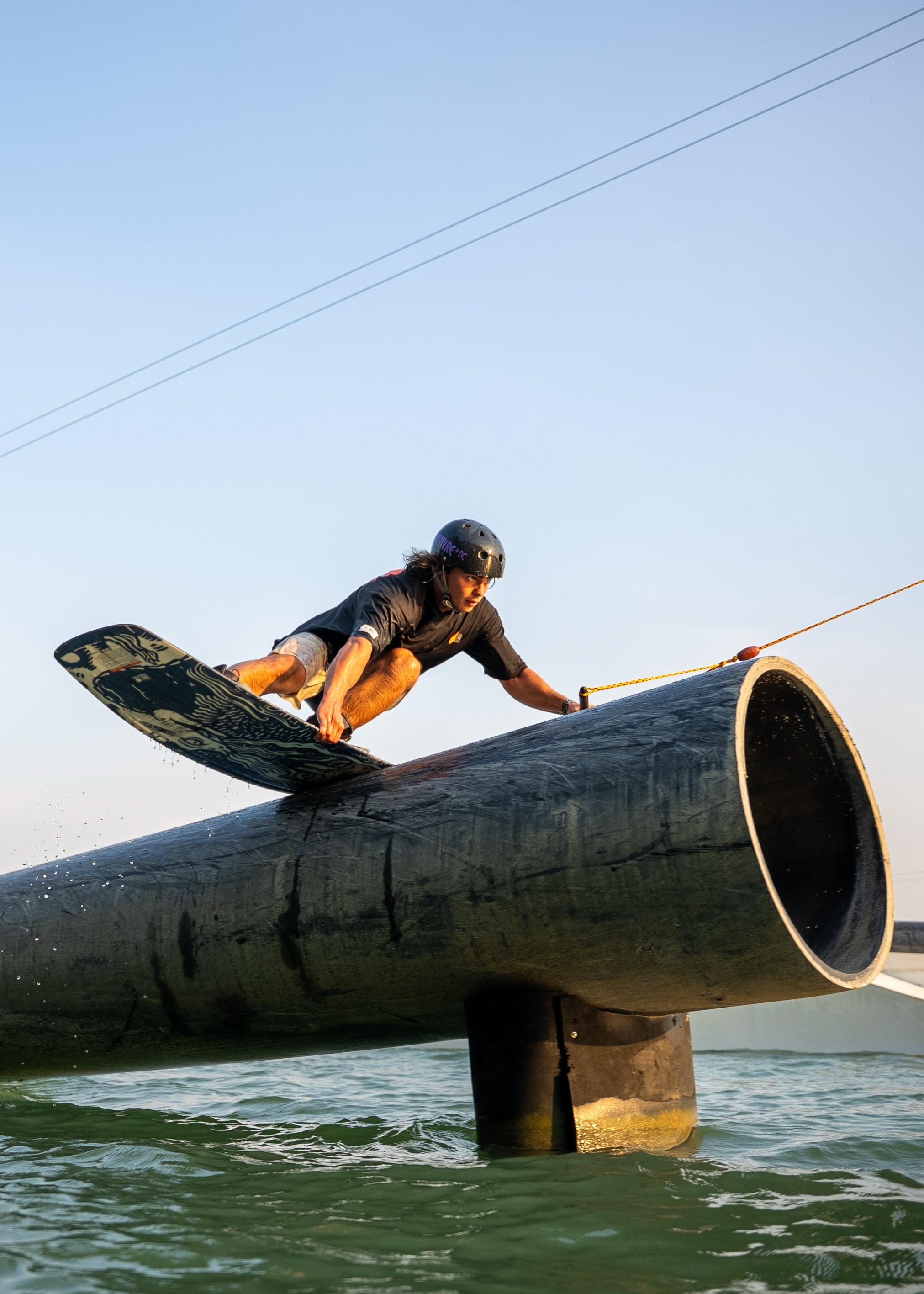 Wakeboarder in helmet gliding over a large pipe above water, holding a rope under clear sky.