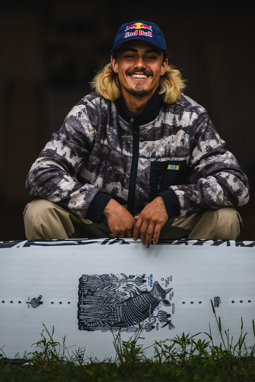 Person smiling, sitting behind a snowboard, wearing a cap and patterned jacket.