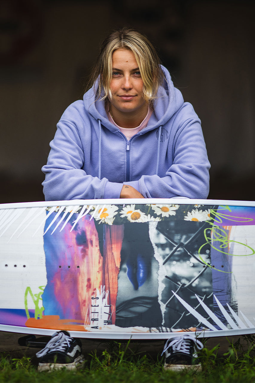 Person in a purple hoodie with blond hair, holding a colorful snowboard with abstract designs.