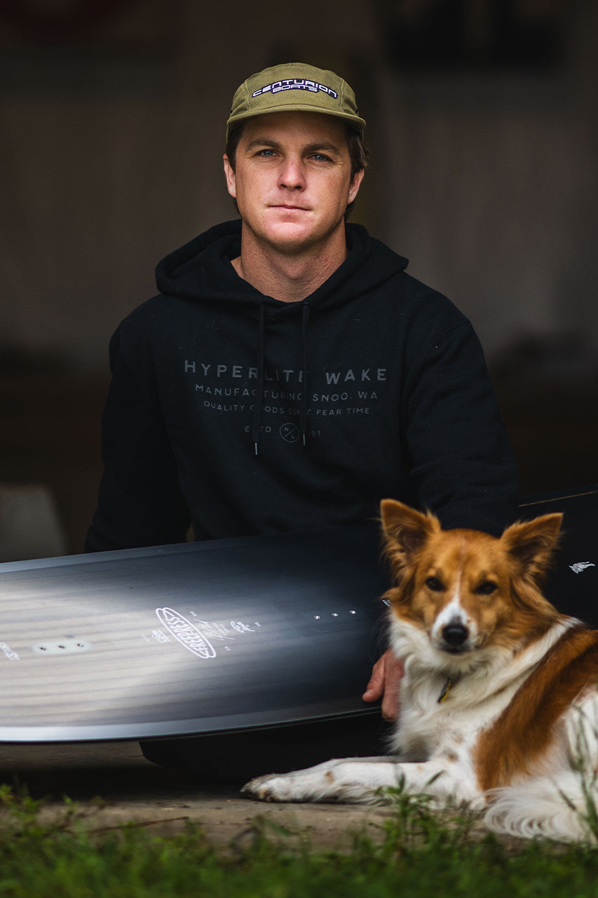 Person in a hoodie and cap with a dog, holding a surfboard upright.