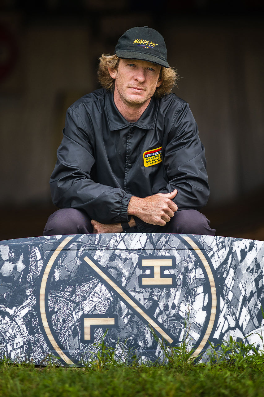Person in cap and jacket posing with a patterned board against a blurred background.