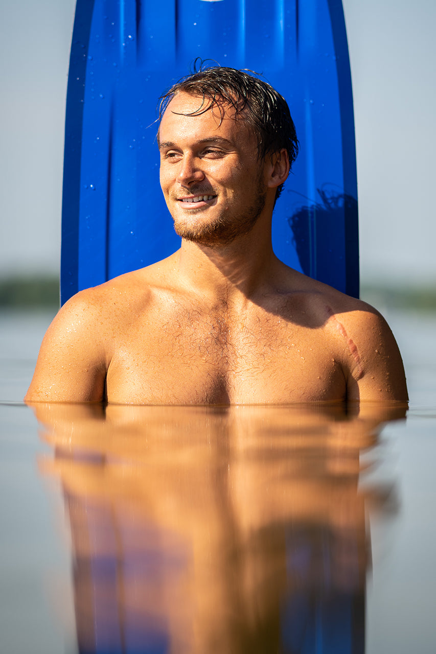 Man standing chest-deep in water, smiling, with a blue surfboard behind him.