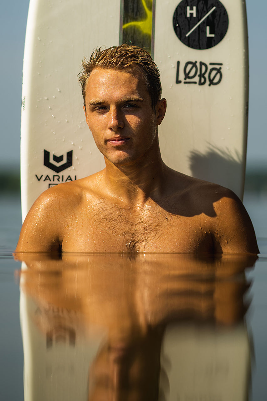 Person chest-deep in water in front of a surfboard, with sunlight highlighting them.