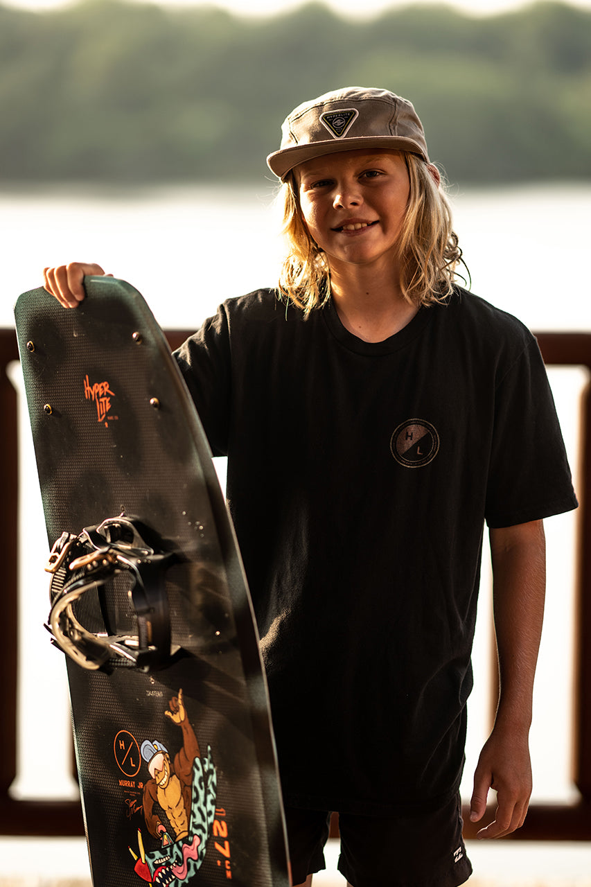Young person with a wakeboard and cap, smiling by a lake.