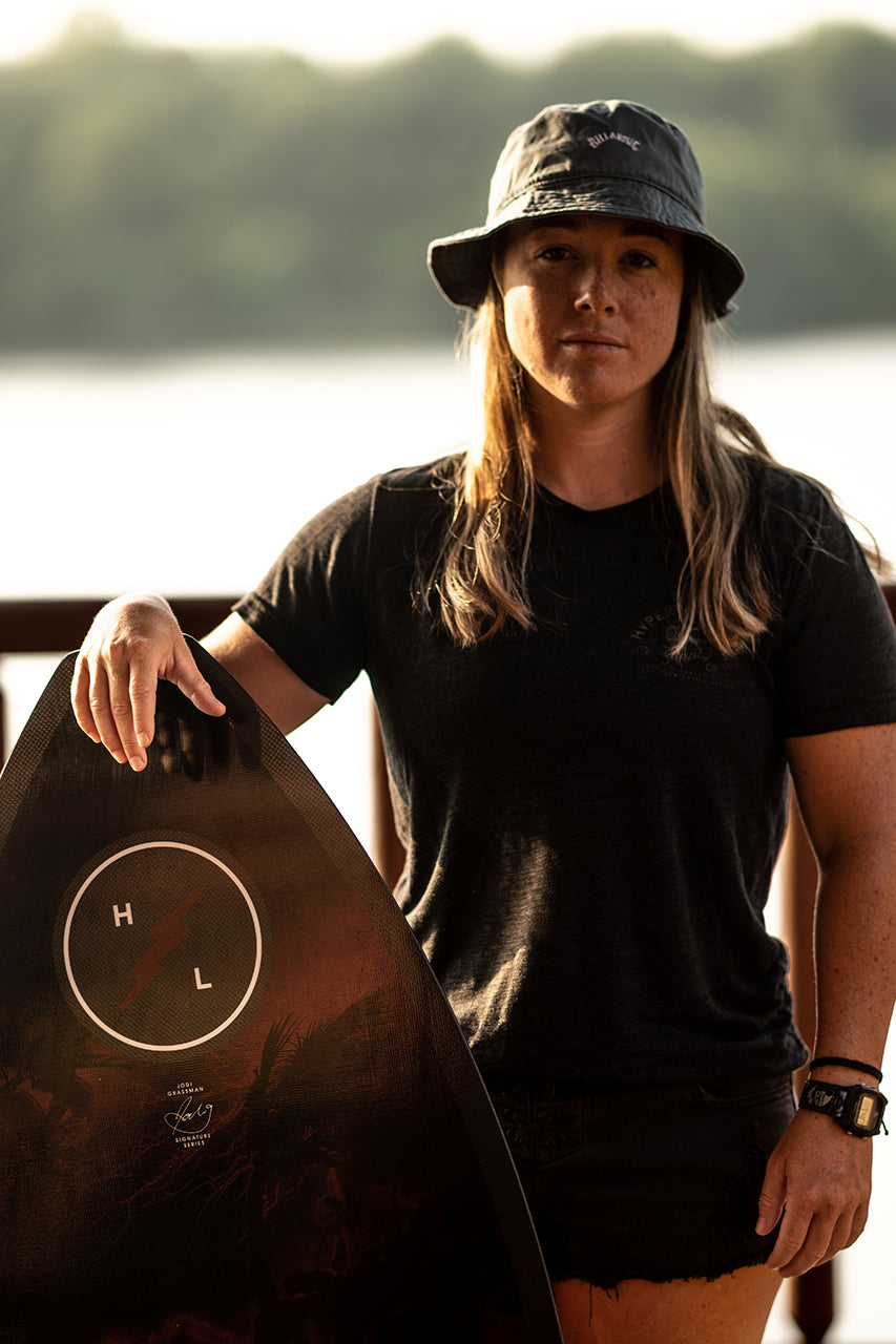 Person holding a surfboard on a deck, wearing a bucket hat and dark clothing.