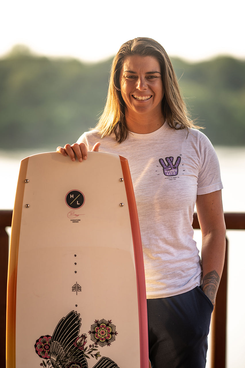 Person smiling and holding a decorated wakeboard, with trees in the background.