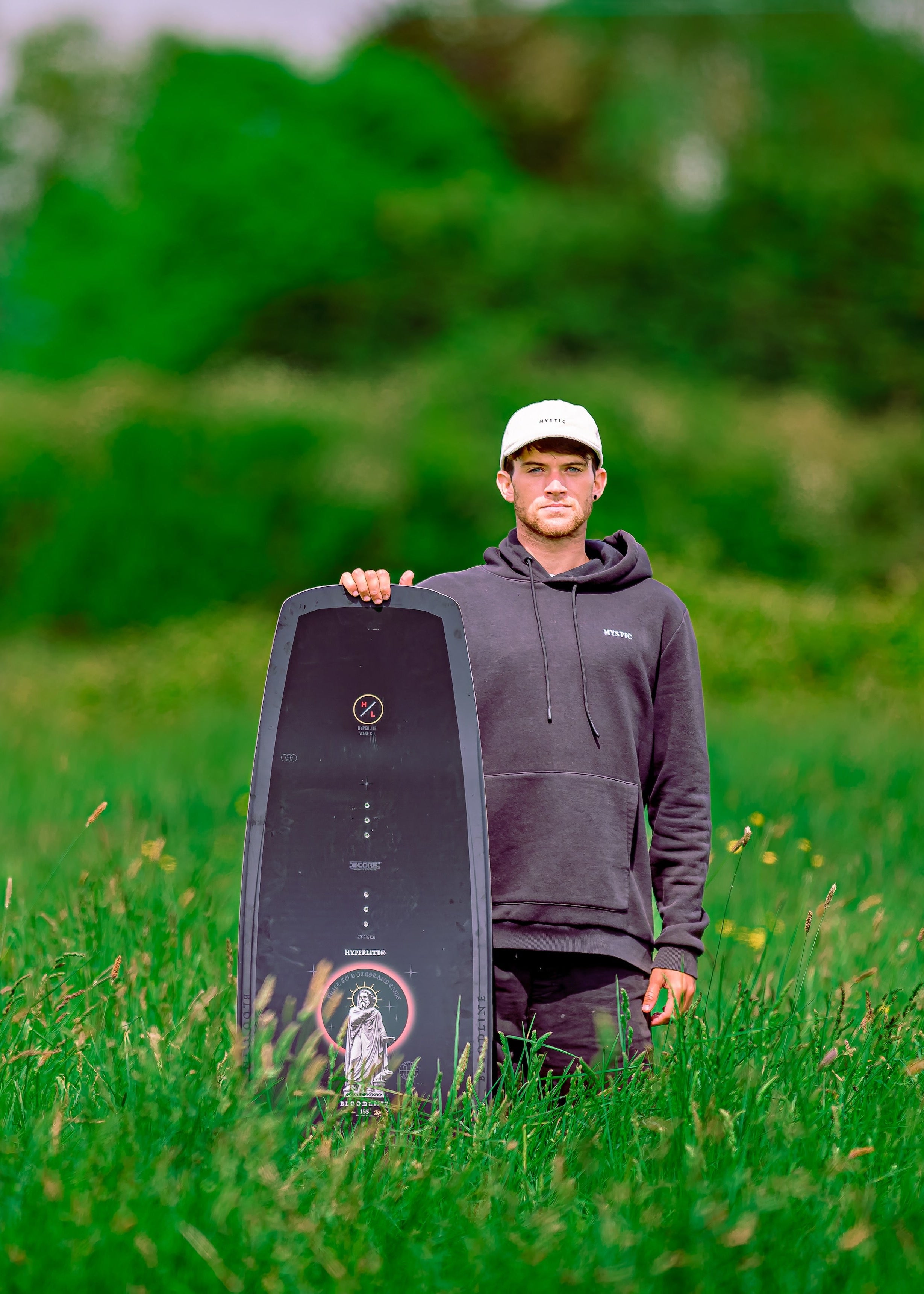 Man standing in tall grass holding a wakeboard upright beside him.
