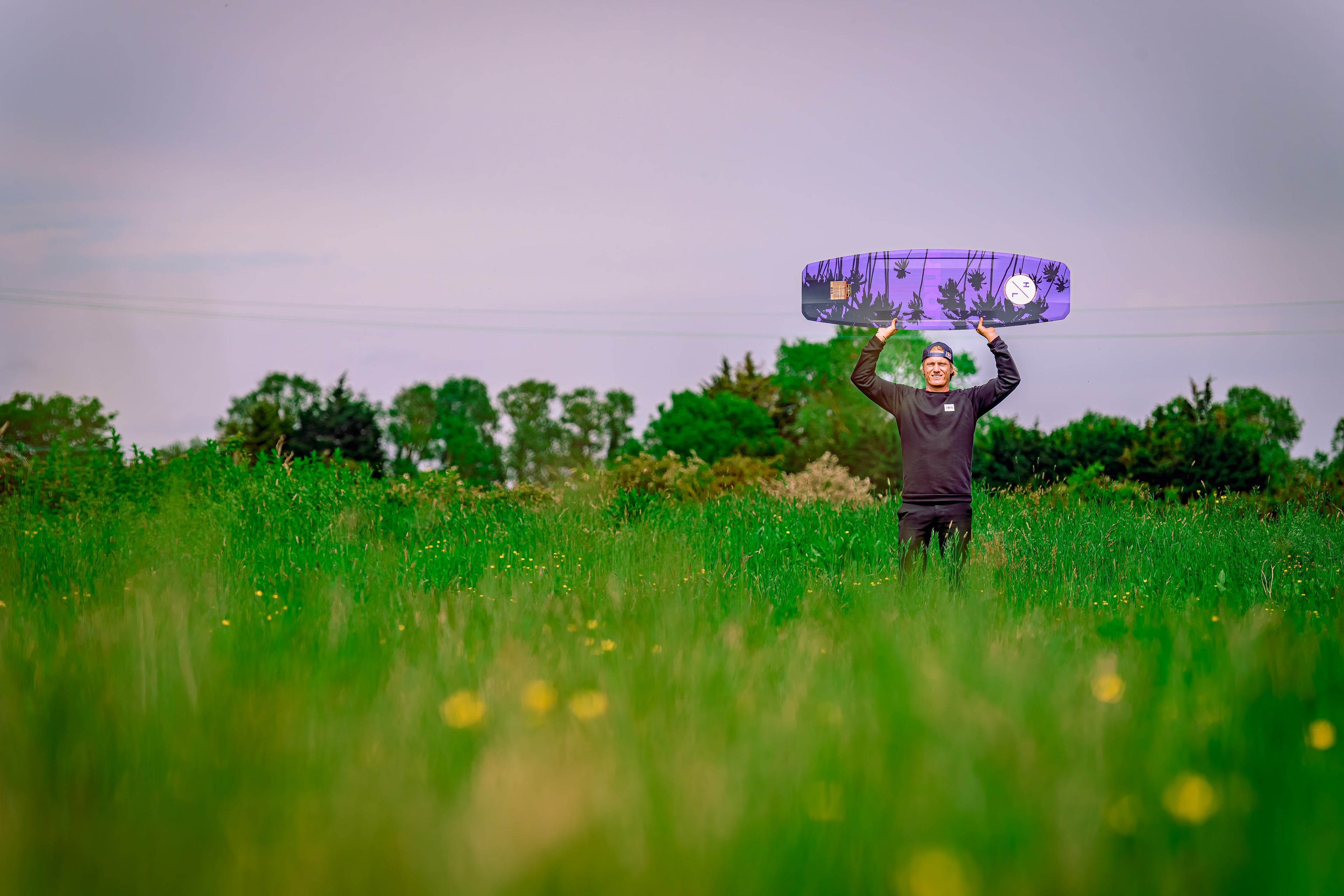Person in a field holding a purple skateboard overhead, surrounded by green grass and trees.