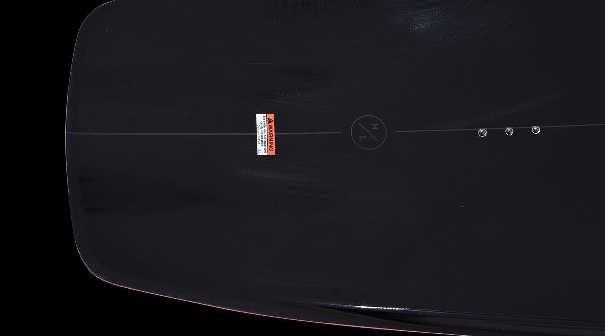 Close-up of mounting inserts and warning label on the black deck of the Pleasure wakeboard.