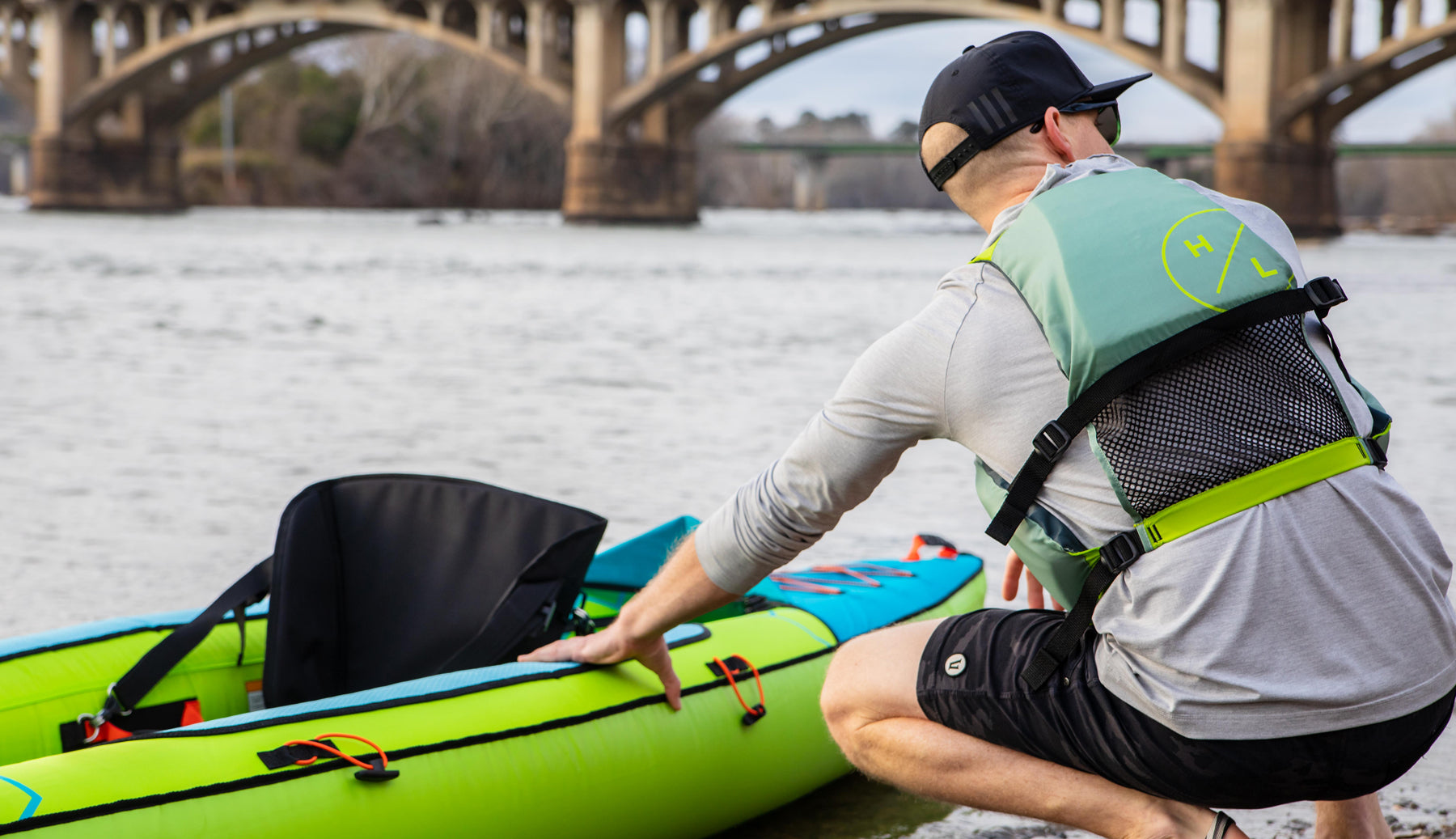 Person preparing a green kayak by a river with a bridge in the background.