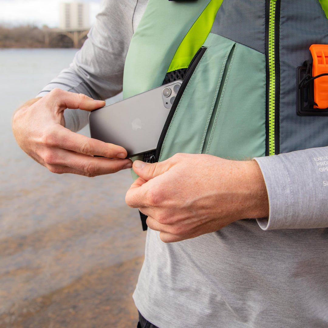 Person putting a smartphone into a life jacket pocket by a body of water.
