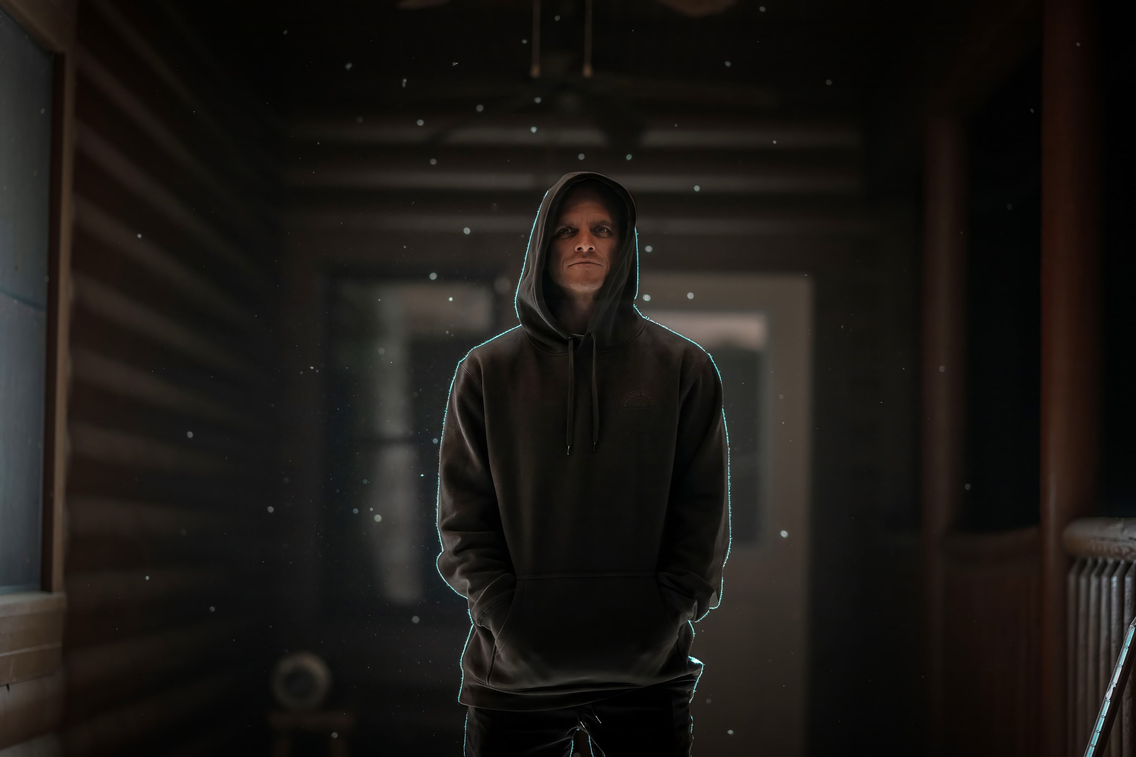 Person in a dark hoodie stands in a dimly lit hallway with specks of light floating around.