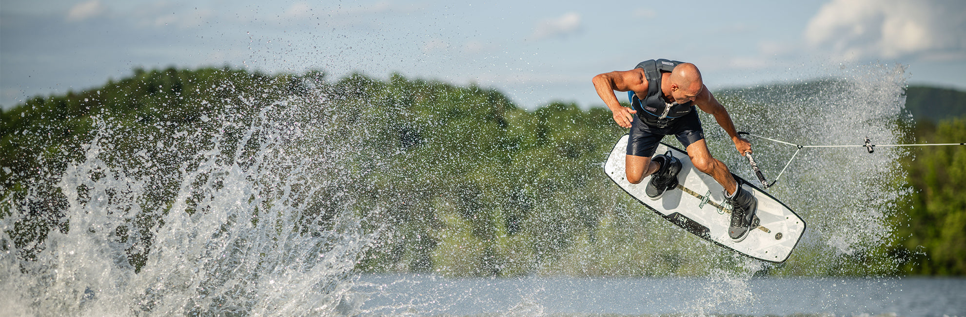 Person wakeboarding, airborne over water with splashes, trees in the background.