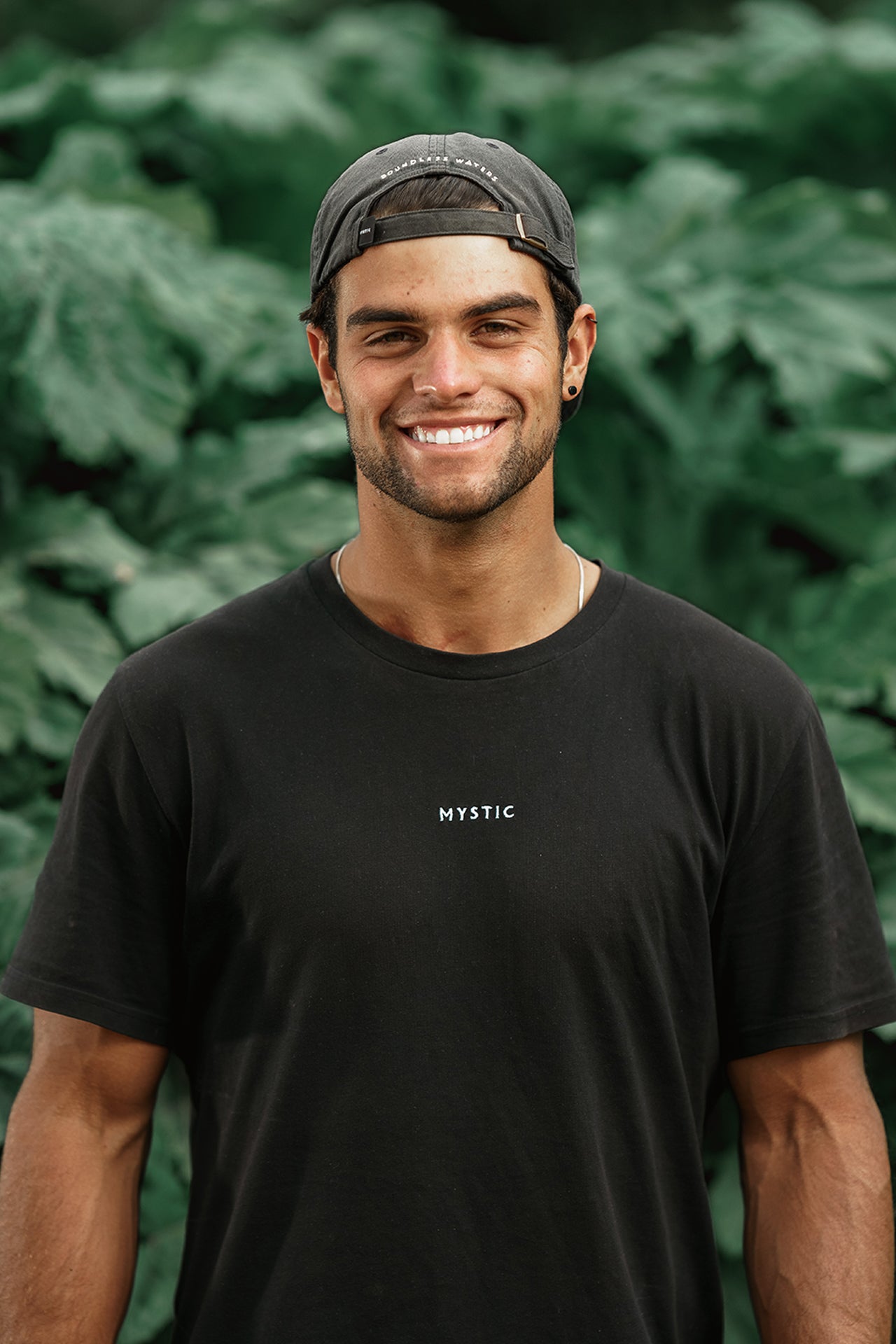 Smiling man in a black t-shirt and cap stands in front of green foliage.
