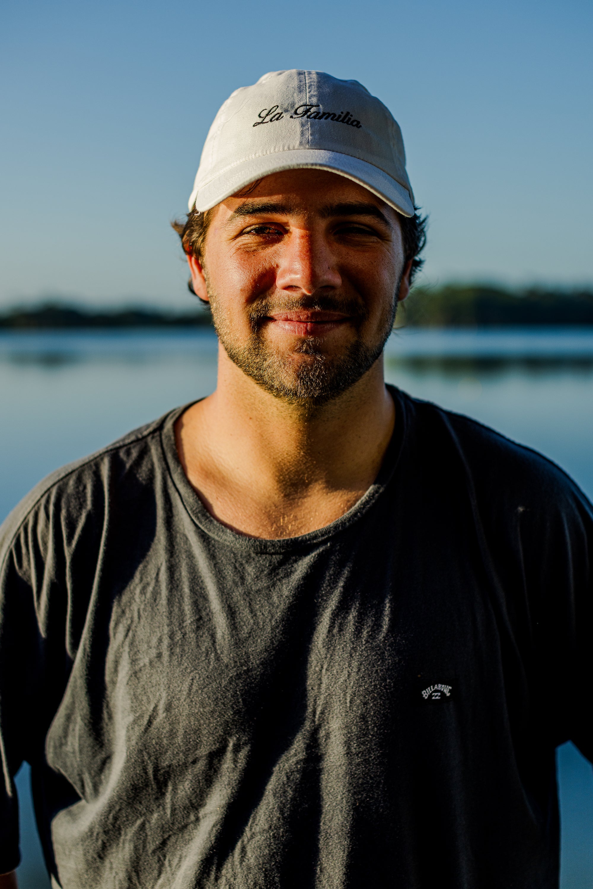 Man in a cap smiling by a lake under a clear blue sky.