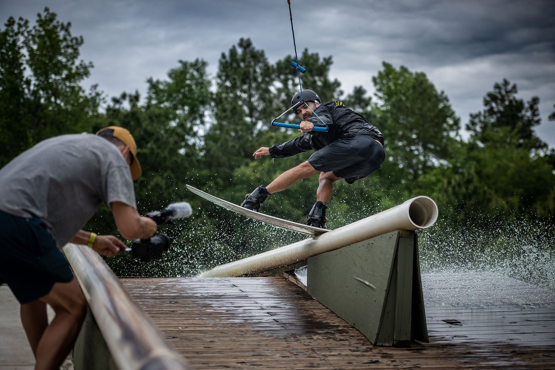 Wakeboarder grinding a rail while being filmed, under a cloudy sky.