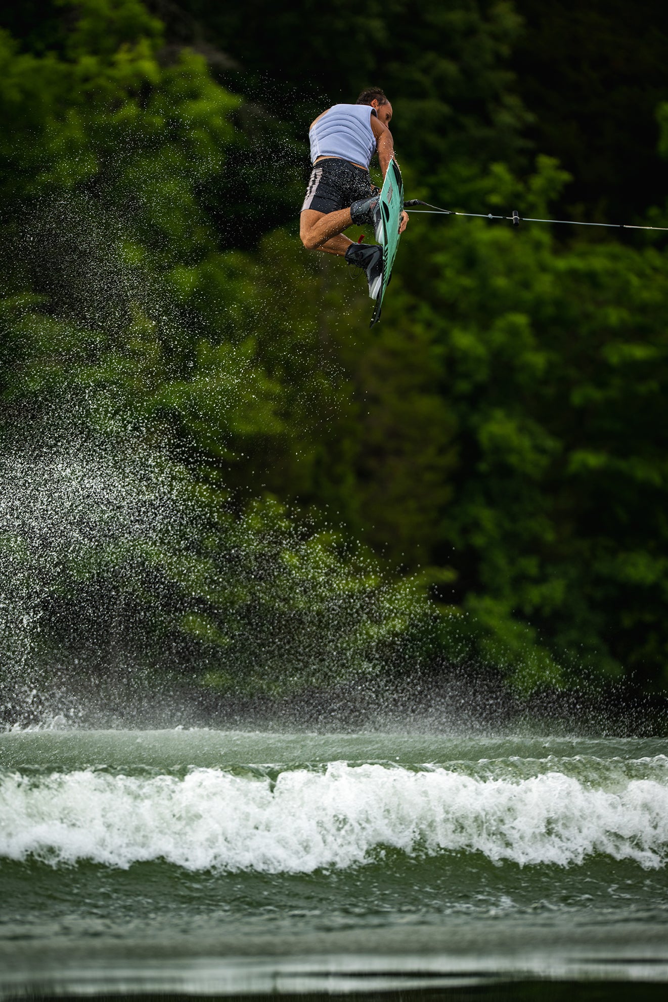 Person wakeboarding, performing a jump over water with lush green trees in the background.