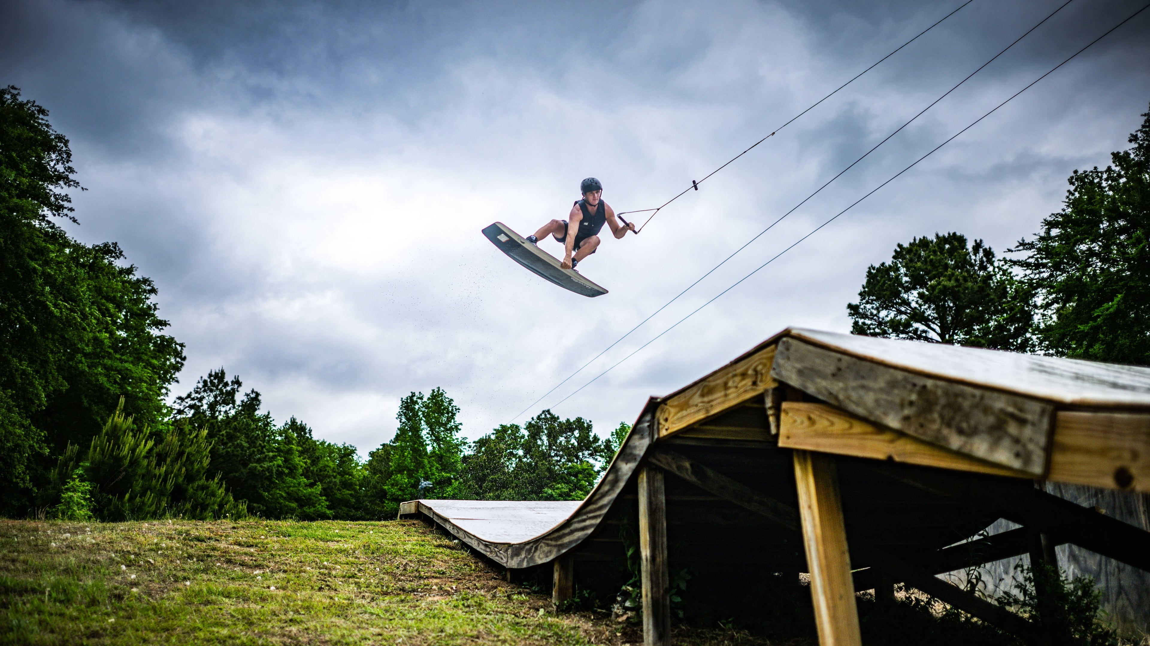 Person wakeboarding mid-air above a wooden ramp on a cloudy day.
