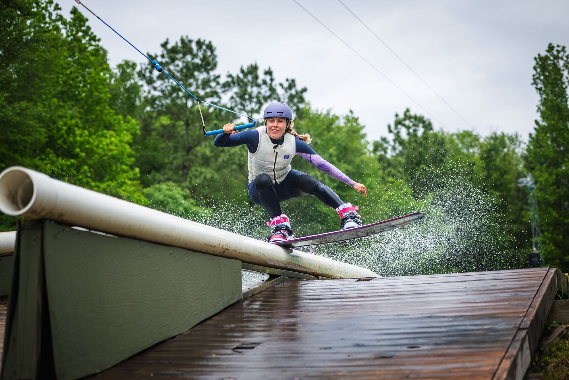 Person wakeboarding on a ramp, wearing a helmet and life vest, surrounded by trees.