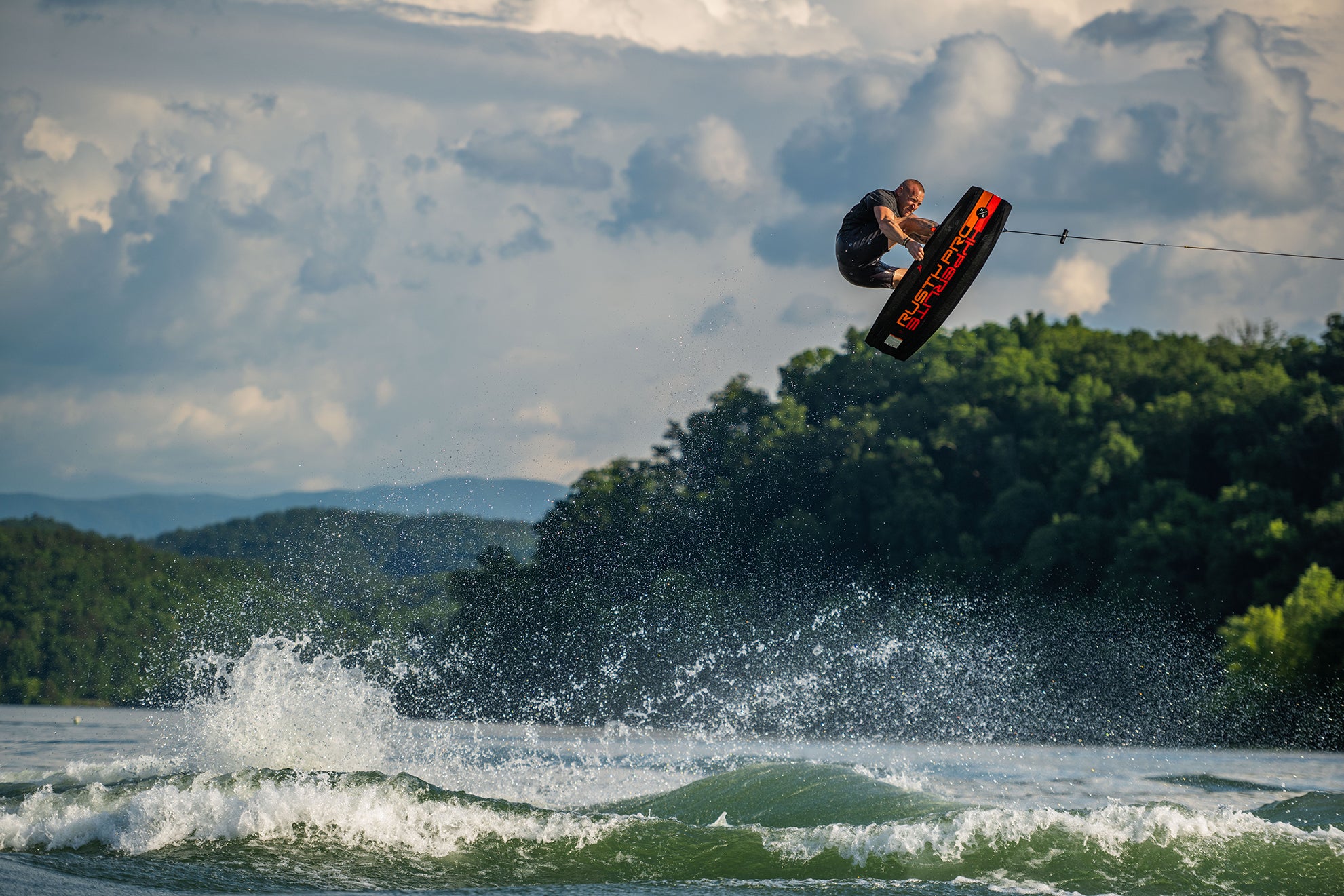 Person wakeboarding mid-air over a lake, with a cloudy sky and forested hills in the background.