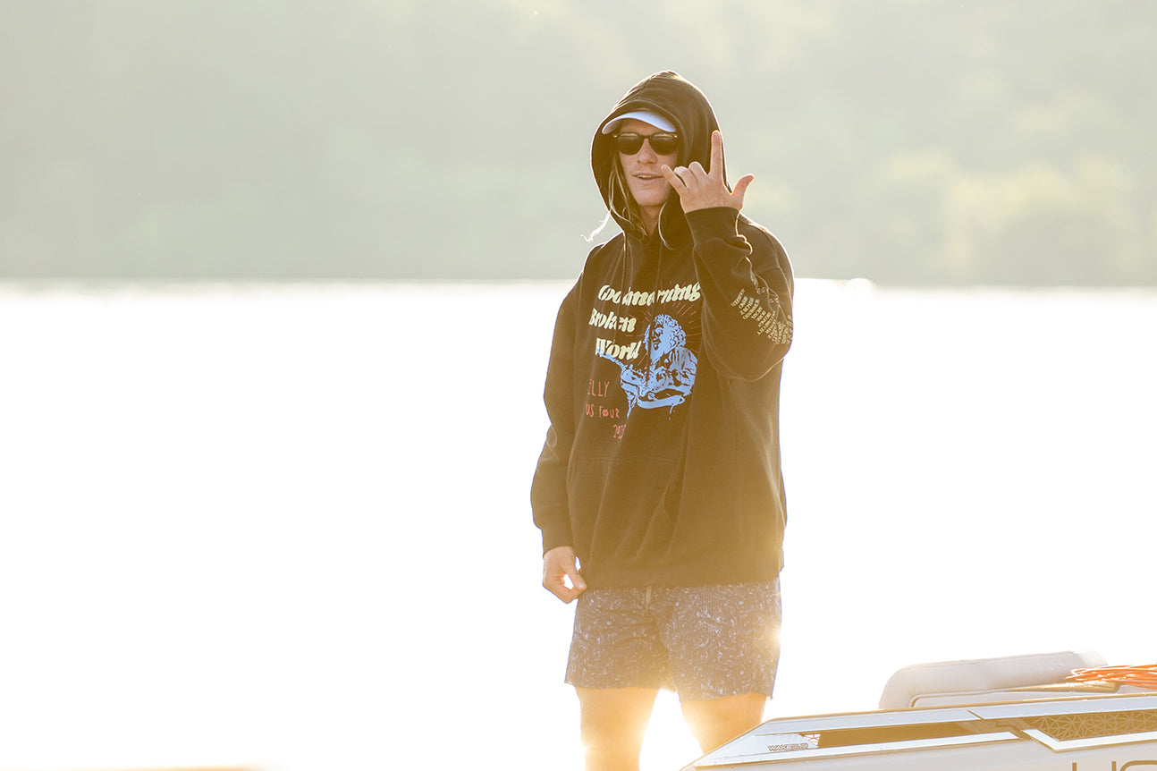 Person in a hoodie and shorts stands outside, making a hand gesture, with a sunny backdrop.