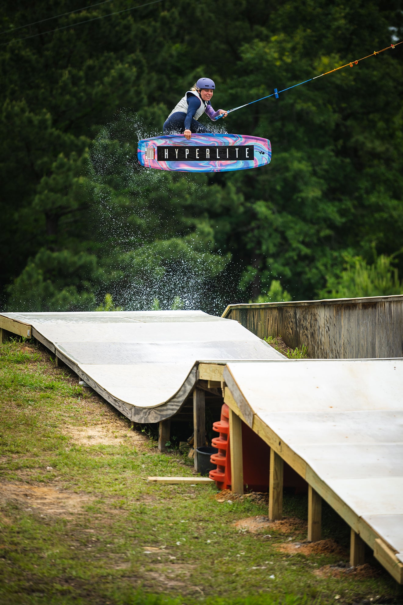 Person wakeboarding over a ramp with green trees in the background.