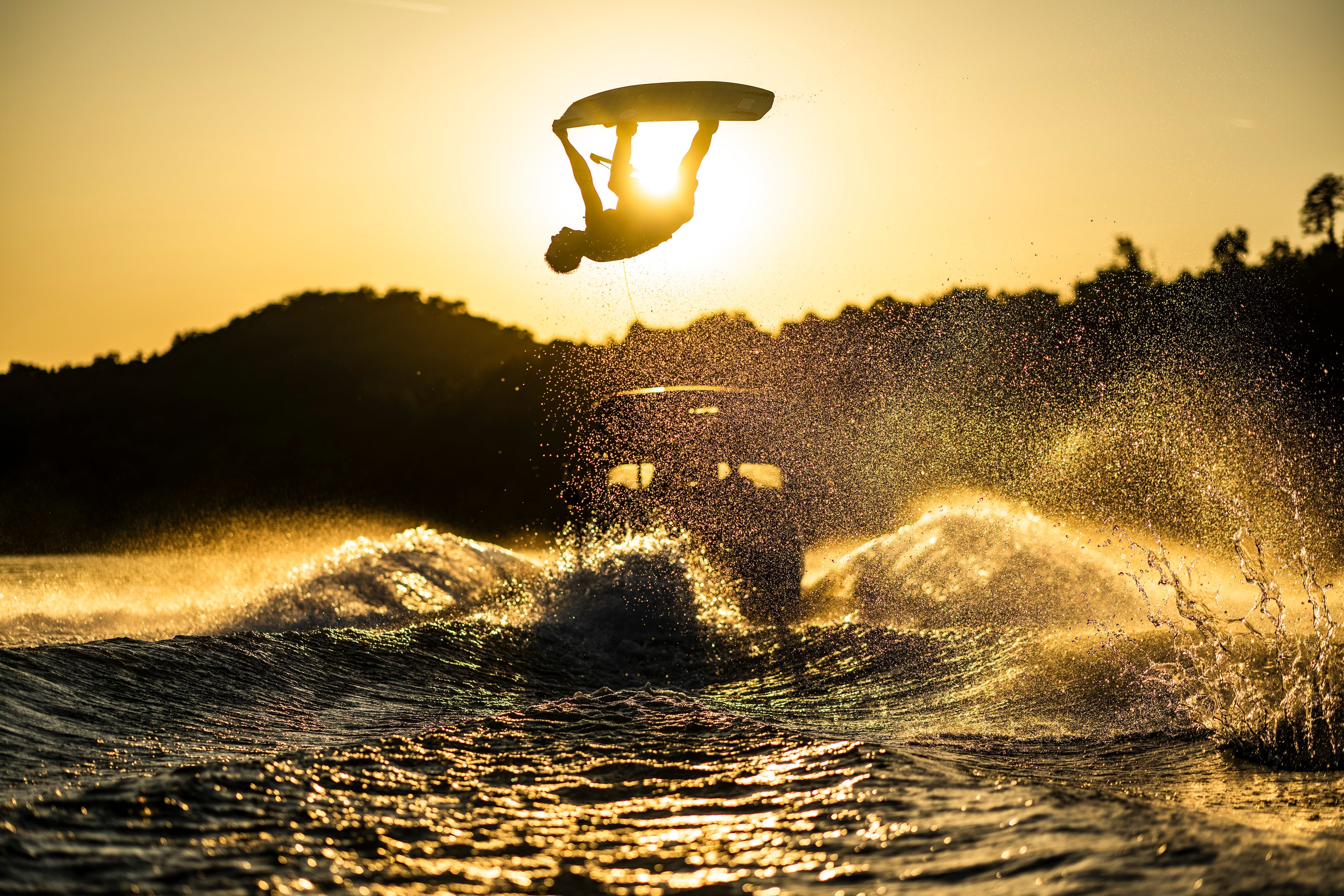 Silhouetted person wakeboarding against the sunset sky, mid-flip above splashing water.