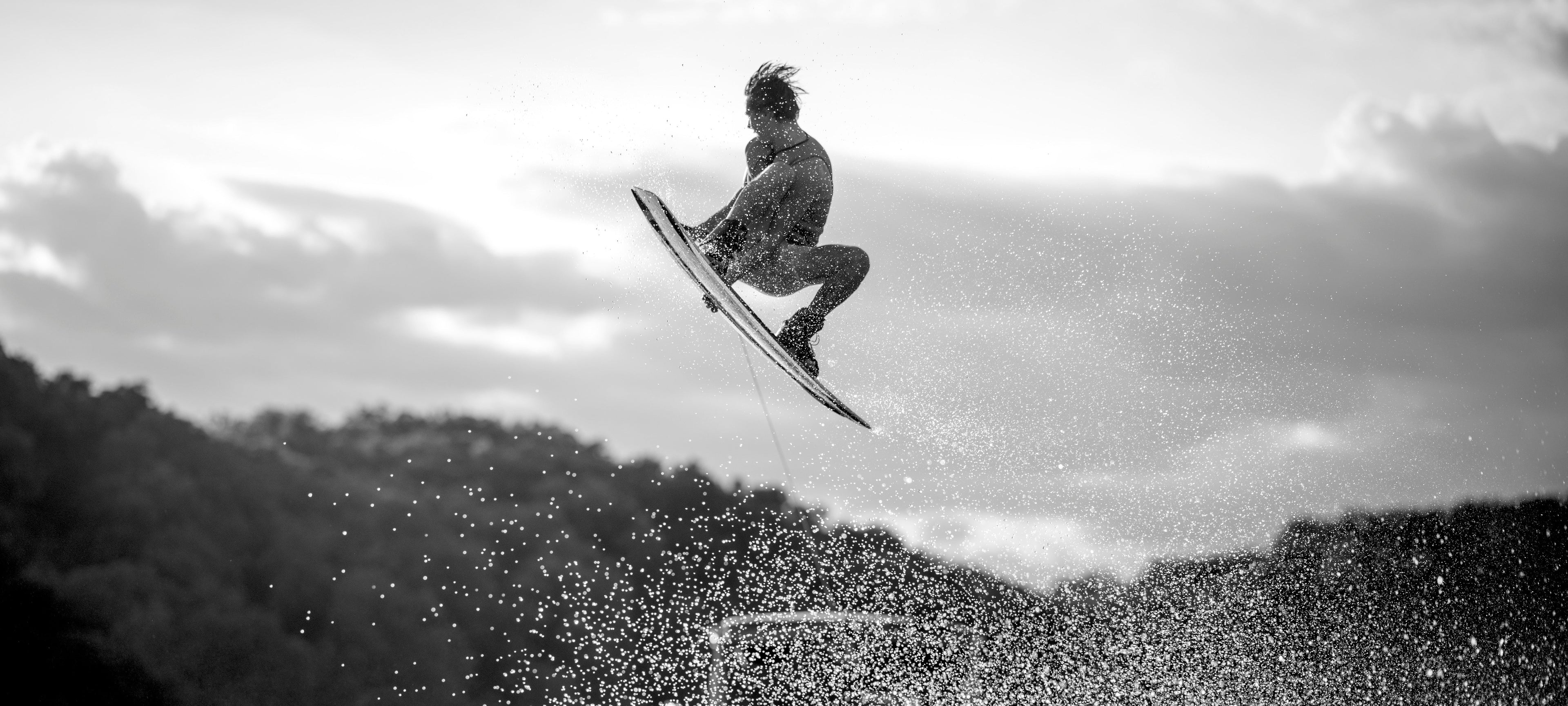 Person wakeboarding in mid-air, silhouetted against a cloudy sky, with water splashing below.