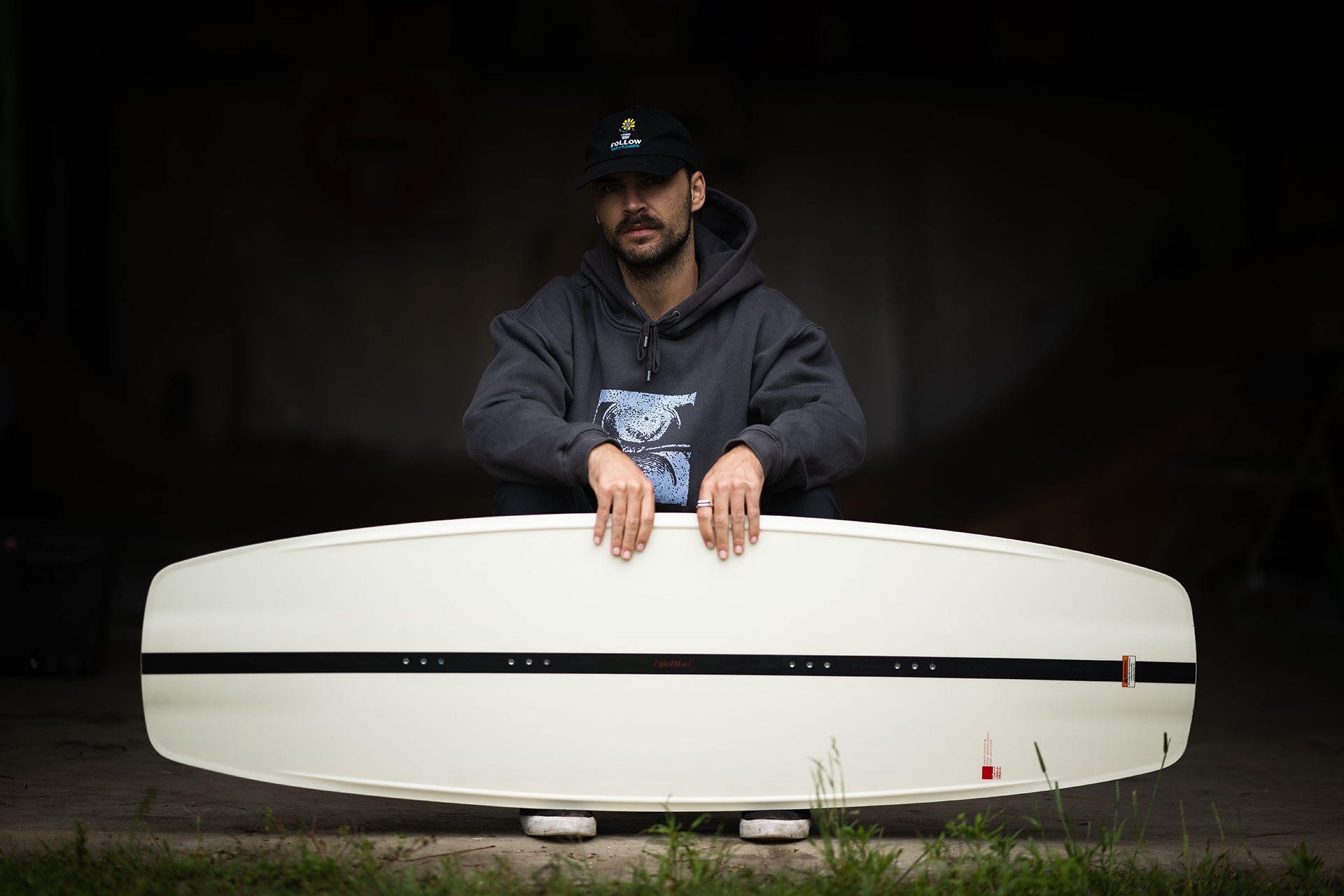 Man in dark hoodie and cap holding a white surfboard.