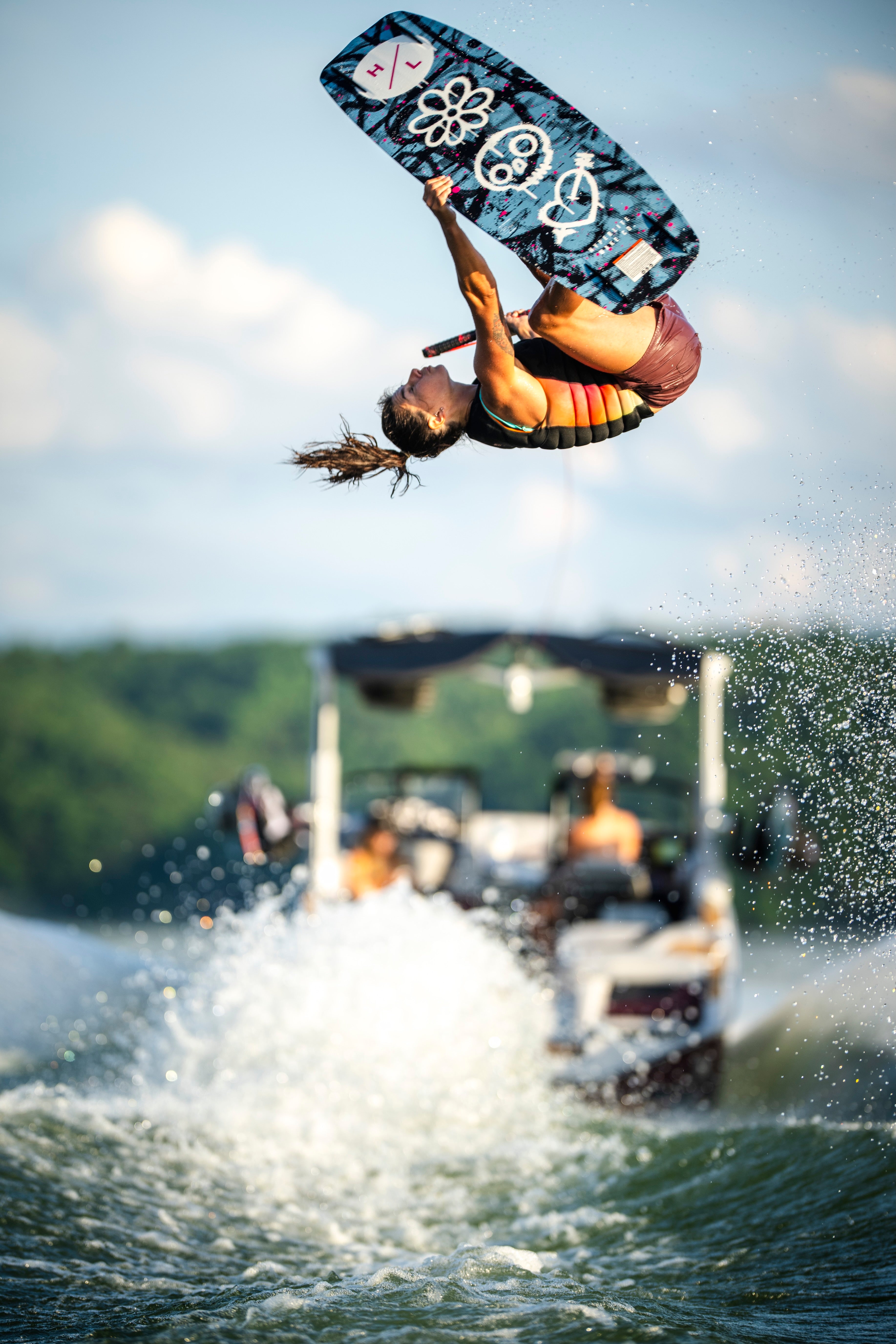 Person performing an aerial trick on a wakeboard, boat and water spray in the background.