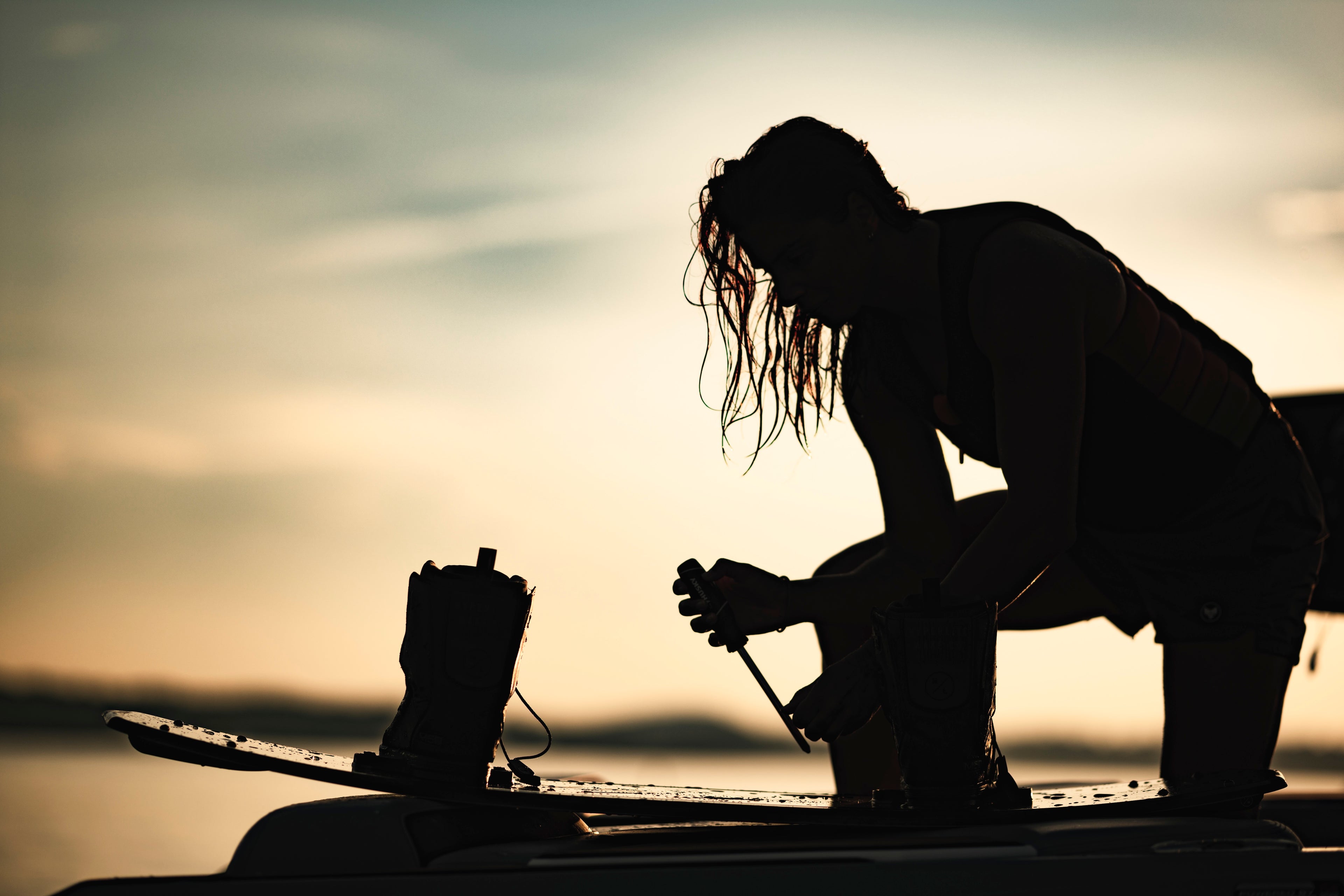 Silhouette of a person adjusting a wakeboard at sunset by the water.